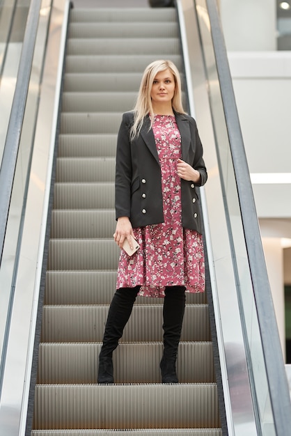 Junge schöne blonde Frau in einem Kleid auf einer Rolltreppe in einem Einkaufszentrum, mit einem Telefon in ihren Händen