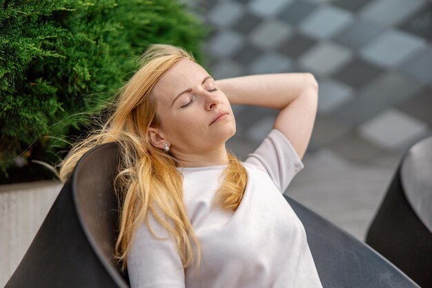 Junge schöne blonde Frau sitzt im Freien auf der hölzernen Terrasse in der Stadt und ruht sich aus