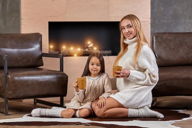 Junge schöne blonde frau umarmt ihre kleine tochter auf der couch mit bechern warmer getränke mutter ca...