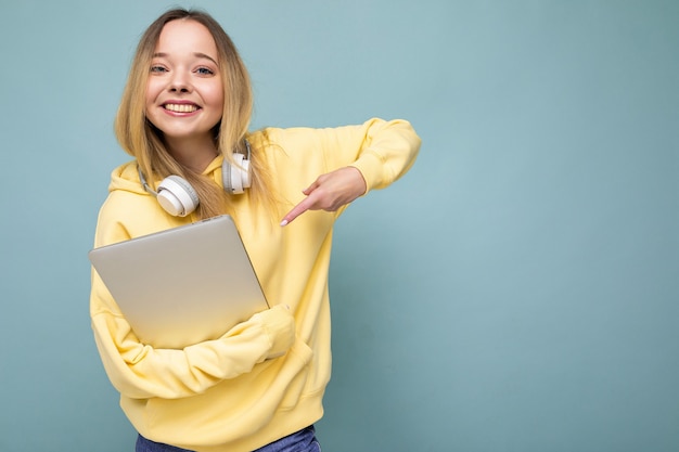 Junge schöne blonde Studentin trägt einen trendigen gelben Hoodie und Kopfhörer mit Computer