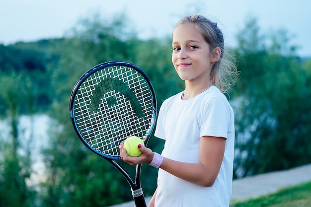 Junge schöne blonde Tennisspielerin kleines Mädchen in Sportbekleidung, weißem T-Shirt und roten Sportshorts, die einen Tennisschläger und einen Ball im Stadion halten. Aktive Tochter spielt am Sommerabend Badminton