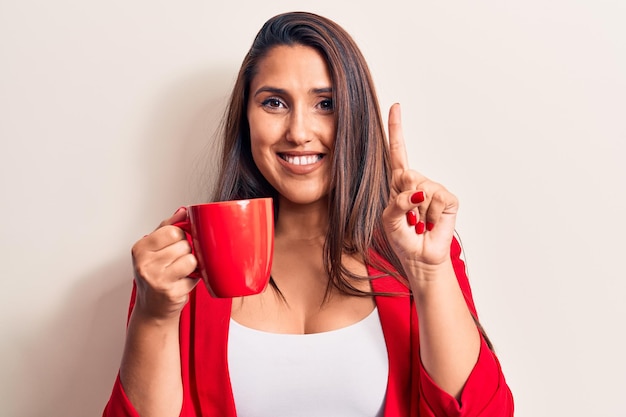 Junge schöne brünette Frau trinkt eine Tasse Kaffee, lächelt mit einer Idee oder Frage und zeigt mit dem Finger und dem glücklichen Gesicht Nummer eins