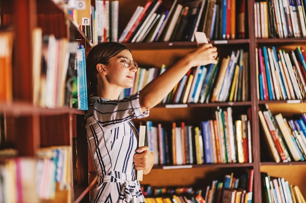 Junge schöne Brünette im Kleid und mit Brillen, die ein Buch halten und Selfie nehmen, während sie in der Bibliothek stehen.