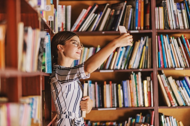 Junge schöne Brünette im Kleid und mit Brillen, die ein Buch halten und Selfie nehmen, während sie in der Bibliothek stehen.