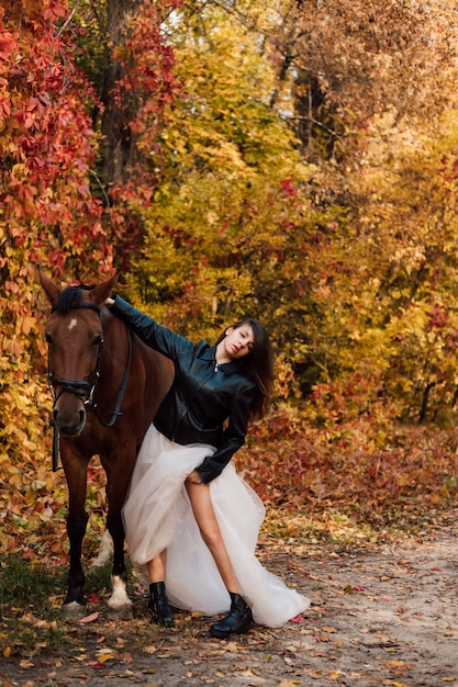 Junge schöne Brünette in einem weißen Kleid und einer schwarzen Lederjacke posiert in der Nähe eines Pferdes