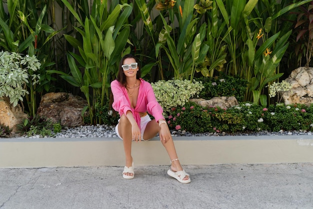 Junge schöne Brünette in einer trendigen Sonnenbrille mit rosafarbenem Hemd, Halsschmuck und Halskette sitzt auf einem Hintergrund aus tropischen Blättern, lacht, lächelt
