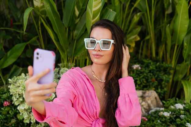 Junge schöne Brünette in einer trendigen Sonnenbrille mit rosafarbenem Hemd, Halsschmuck und Halskette sitzt auf einem Hintergrund aus tropischen Blättern und macht ein Selfie am Telefon