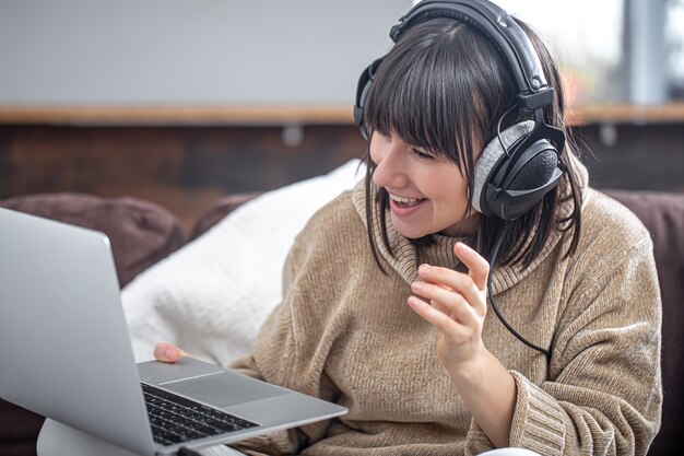 Junge schöne Brünette in Kopfhörern lächelt in einem gemütlichen Pullover und schaut auf den Laptop-Bildschirm. Online-Training, Fernarbeit, Videokommunikation.