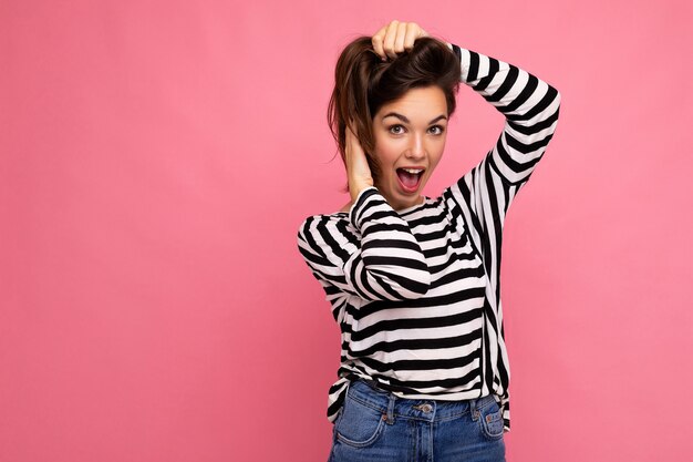 Junge schöne Brunetfrau. Trendige Dame im lässig gestreiften Longsleeve. Positives Weibchen zeigt aufrichtige Gesichtsgefühle. Lustiges Modell auf rosa Hintergrund mit freiem Speicherplatz isoliert.