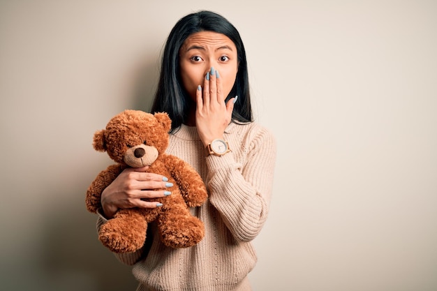 Junge schöne Chinesin mit Teddybär, der über isoliertem weißem Hintergrund steht, bedecken den Mund mit der Hand, die vor Scham wegen des Fehlerausdrucks der Angst schockiert ist, die im geheimen Konzept der Stille Angst hat