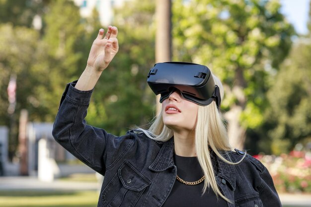 Junge schöne Dame, die eine VR-Brille trägt und ihre Hand im Park in der Luft hält