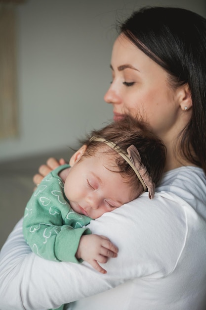 Junge schöne dunkelhaarige Mutter hält ihre neugeborene Tochter in ihren Armen in einem gemütlichen Zuhause Familienporträt Mutterschaft Junge Frau küsst ihr 2 Monate altes Baby