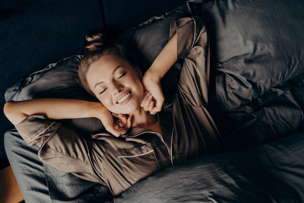 Junge schöne entspannte brünette Frau im Satinpyjama, die sich im Bett ausdehnt