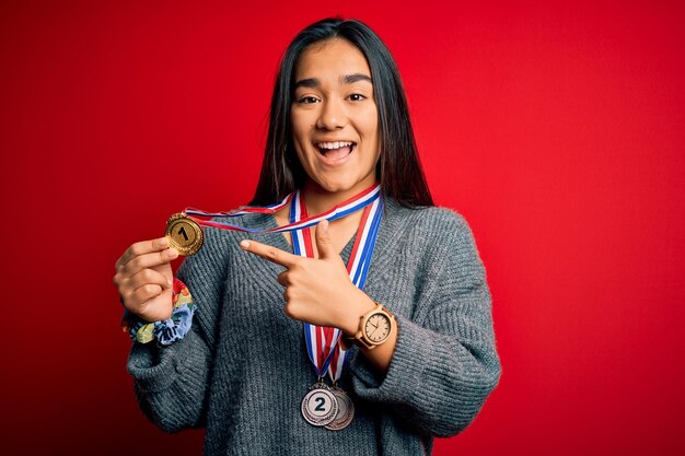 Foto junge, schöne, erfolgreiche asiatische frau mit medaillen steht über einem roten hintergrund und zeigt sehr glücklich mit hand und finger