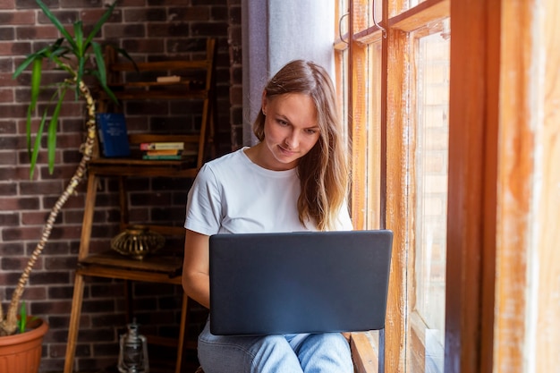 Junge schöne Frau, die am Fenster sitzt und mit Laptop arbeitet.