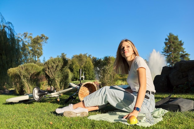 Junge schöne Frau, die auf dem Gras im Park sitzt, Studentin mit Notizbuch, Rucksack, Fahrrad