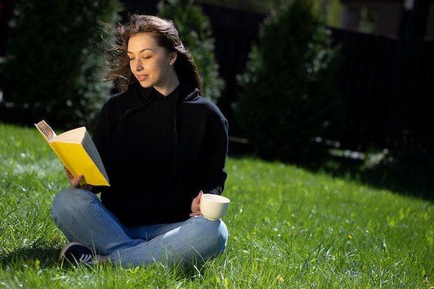 Junge schöne Frau, die auf Gras im Park mit Papierbuch und Tasse Tee sitzt, die ruht