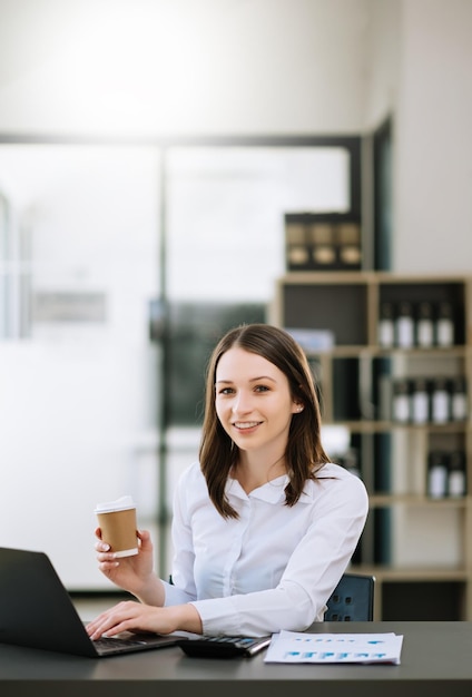 Junge schöne Frau, die auf Tablet und Laptop tippt, während sie am modernen OfficexA des Arbeitsholztisches sitzt