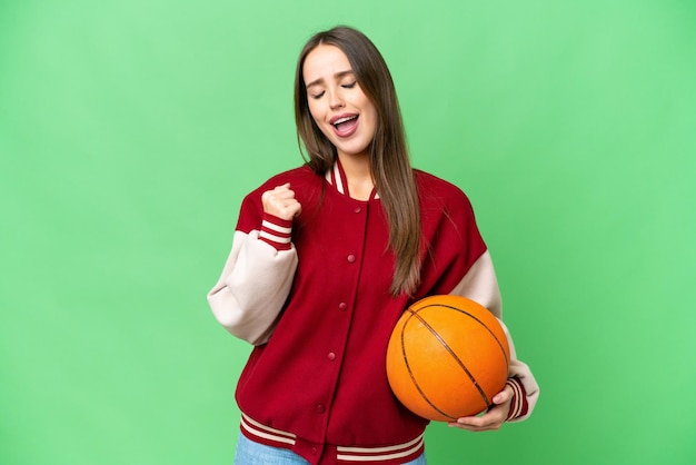 Junge schöne Frau, die Basketball über isoliertem Chroma-Key-Hintergrund spielt und einen Sieg feiert
