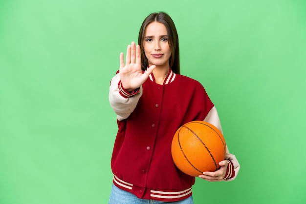 Junge schöne Frau, die Basketball über isoliertem Chroma-Key-Hintergrund spielt und Stoppgeste macht