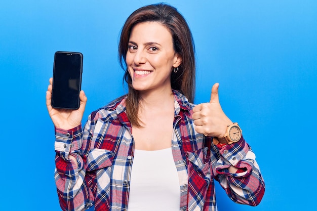 Junge schöne Frau, die ein Smartphone hält und einen Bildschirm zeigt, der glücklich und positiv lächelt, den Daumen hoch macht und ein Genehmigungszeichen macht