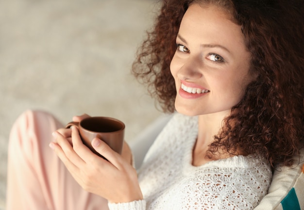 Junge schöne Frau, die eine Tasse Kaffee hält und auf dem Sofa im Zimmer sitzt