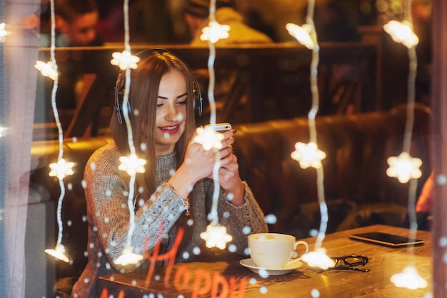 Junge schöne Frau, die im Kaffee, trinkender Kaffee sitzt. Vorbildliches Hören von Musik. Weihnachten, Neujahr, Valentinstag, Winterferien