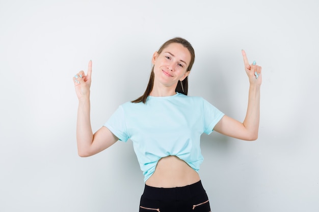 Junge schöne Frau, die im T-Shirt, in der Hose nach oben zeigt und glückselig aussieht, Vorderansicht.