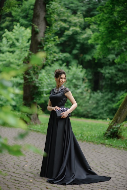 Junge schöne Frau, die in einem schwarzen Kleid in einem Park aufwirft. Eine Handtasche in den Händen halten.