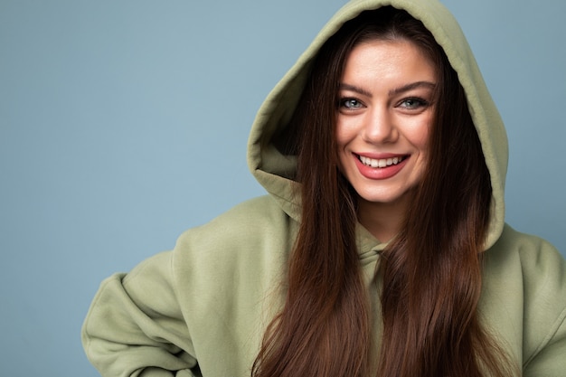 Junge schöne frau, die kamera betrachtet. trendiges mädchen in lässiger hipster-hoodie-kleidung. positives weibchen zeigt gesichtsgefühle. lustiges modell auf blauem hintergrund mit freiem speicherplatz isoliert.