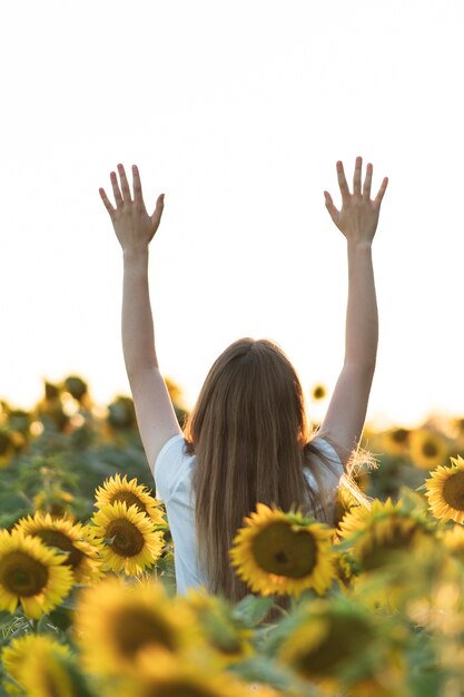 Junge schöne Frau, die lächelt und Spaß in einem Sonnenblumenfeld an einem schönen Sommertag mit erhobenen Armen hat.