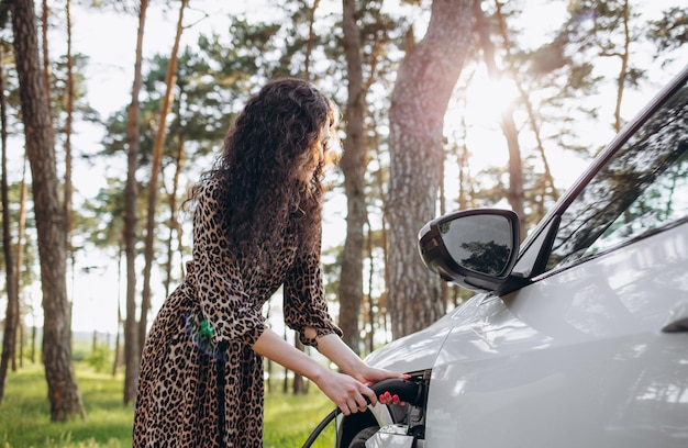 Junge schöne Frau, die mit einem Elektroauto unterwegs ist und an einer Ladestation Halt macht, steht am Kabel, surft im Internet auf dem Smartphone und lächelt fröhlich, während sie sich auflädt