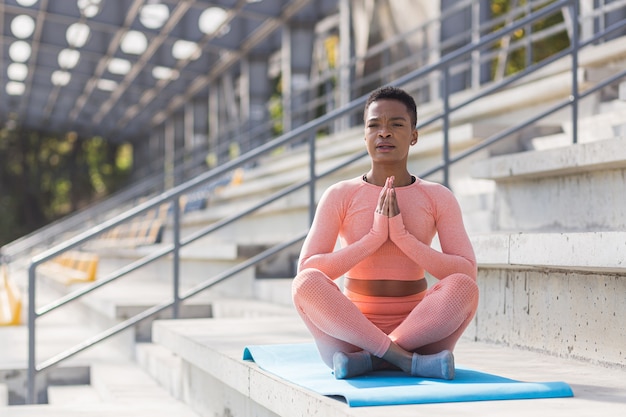Junge schöne Frau, die morgens meditiert, Afroamerikanerin in einem rosa Sportanzug führt Fitnessübungen mit Atmung in der Nähe des Stadions durch