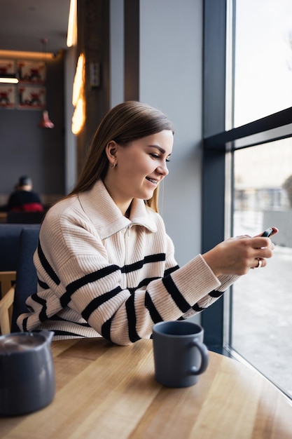 Junge schöne Frau, die Smartphone hält und betrachtet, während sie in der Cafeteria sitzt. Glücklicher Universitätsstudent, der Handy verwendet. Geschäftsfrau trinkt Kaffee und lächelt.
