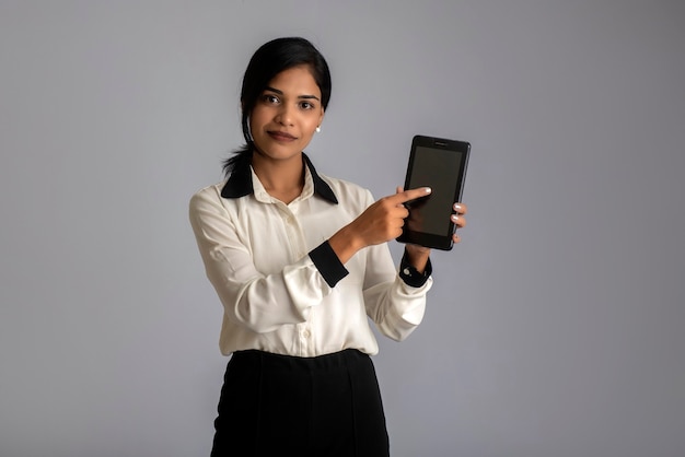 Junge schöne Frau, die Smartphone oder Handy oder Tablet-Telefon des leeren Bildschirms auf einer grauen Wand hält und zeigt.