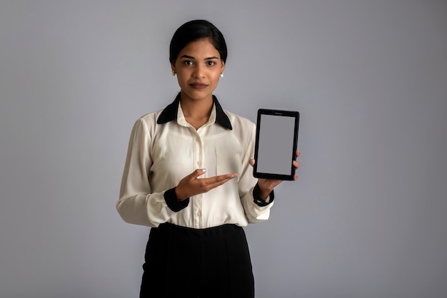 Junge schöne Frau, die Smartphone oder Handy oder Tablet-Telefon des leeren Bildschirms auf einer grauen Wand hält und zeigt.