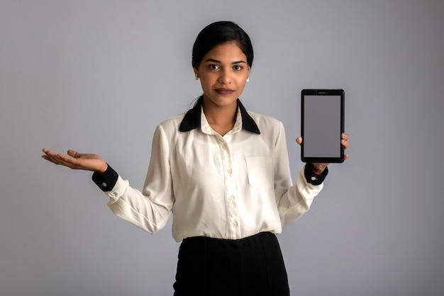 Junge schöne Frau, die Smartphone oder Handy oder Tablet-Telefon des leeren Bildschirms auf einer grauen Wand hält und zeigt.