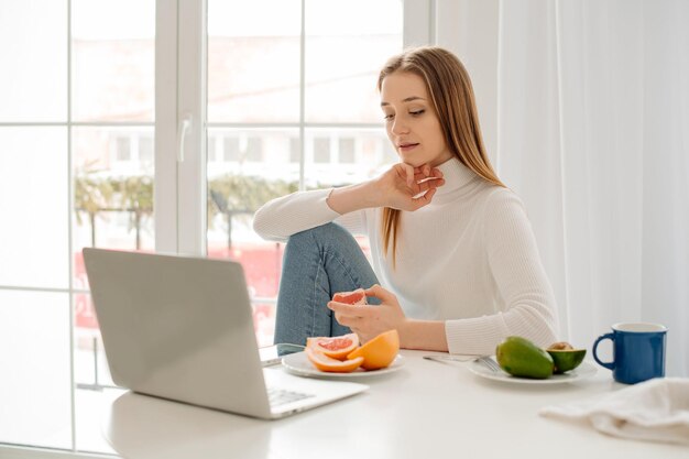 Junge schöne Frau, die zu Hause mit Laptop vor einem großen Fenster mit Morgenlicht arbeitet