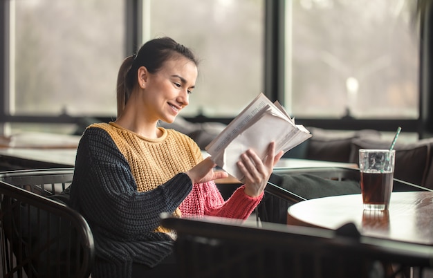 Junge schöne Frau im orangefarbenen Pullover, der interessantes Buch im Café liest