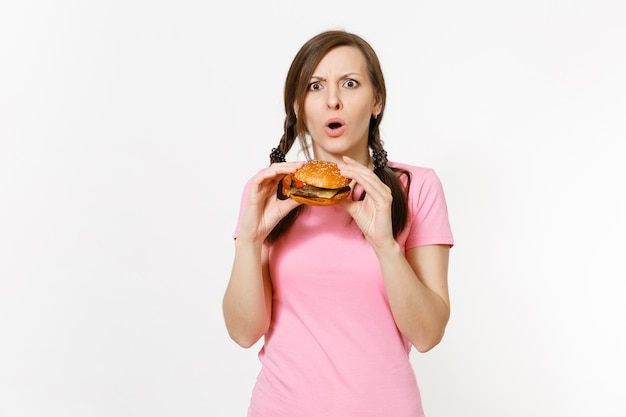 Junge schöne Frau im rosa T-Shirt mit Zöpfen, die in den Händen halten Burger isoliert auf weißem Hintergrund. Richtige Ernährung oder amerikanisches klassisches Fast Food. Kopieren Sie Platz für Werbung. Werbefläche.