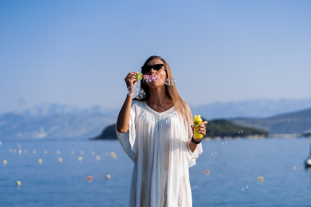 Junge schöne Frau im weißen Kleid und in der Sonnenbrille, die Seifenblasen auf Pier mit Meerblickhintergrund bläst. Das Konzept von Freude, Leichtigkeit und Freiheit im Urlaub. Das Mädchen genießt den Rest.