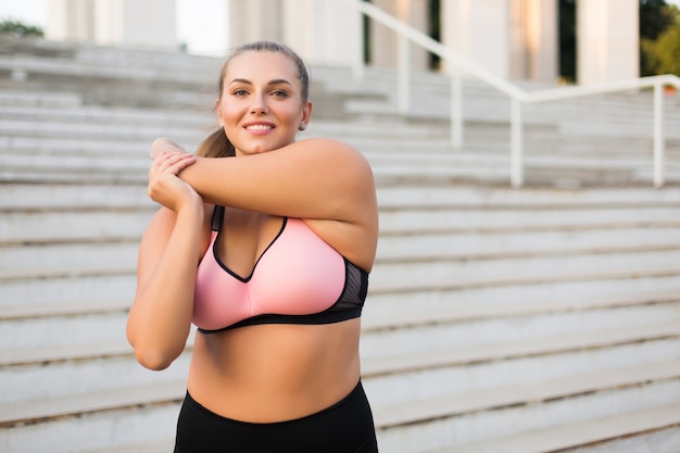 Junge schöne Frau in Übergröße in rosa sportlichem Oberteil und Leggings, die sich freudig dehnen, während Zeit im Freien verbringen