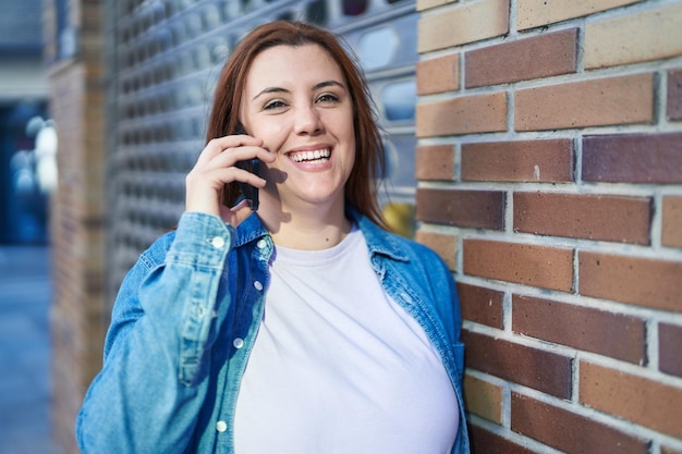 Junge schöne Frau in Übergröße lächelt selbstbewusst und spricht auf der Straße auf dem Smartphone