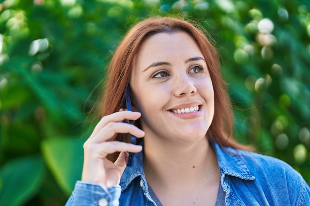 Junge schöne Frau in Übergröße lächelt selbstbewusst und spricht im Park auf dem Smartphone