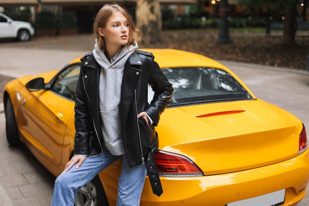 Junge schöne Frau in der Lederjacke, die sich auf gelben Sportwagen stützt, während nachdenklich