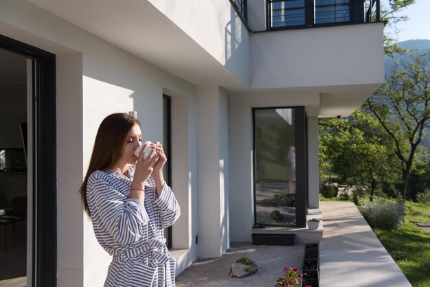 junge schöne frau in einem bademantel, die morgens kaffee vor ihrer luxusvilla genießt