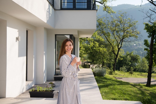 junge schöne frau in einem bademantel, die morgens kaffee vor ihrer luxusvilla genießt