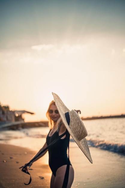 Junge schöne Frau in einem schwarzen Badeanzug und Hut mit Brille geht bei Sonnenuntergang am Strand entlang Das Konzept der Erholung am Meer Selektiver Fokus