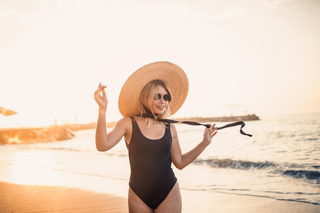 Junge schöne Frau in einem schwarzen Badeanzug und Hut mit Brille geht bei Sonnenuntergang am Strand entlang Das Konzept der Erholung am Meer Selektiver Fokus