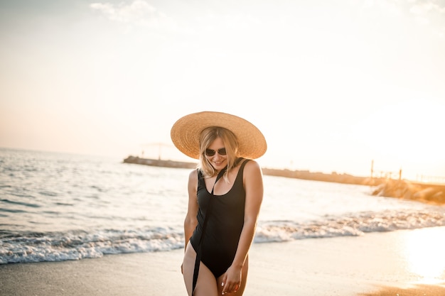 Junge schöne Frau in einem schwarzen Badeanzug und Hut mit Brille geht bei Sonnenuntergang am Strand in der Türkei entlang. Das Konzept der Erholung am Meer. Selektiver Fokus